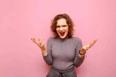 Expressive cute girl is angry on a pastel pink background, yelling and looking at the camera with her hands up. Emotional lady with curly hair and stylish casual clothes scream with anger.