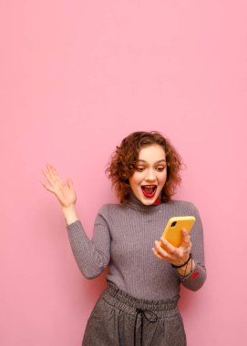 Extremely stylish lady with curly red hair and a smartphone in her hands rejoices and looks insane at the screen of a smartphone. Shocked girl looks at smartphone and emotion rejoices. Vertical photo
