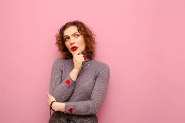 Attractive curly lady in casual clothes isolated on pastel pink background, looks away with pensive face on copy space. Pensive girl in sweater with hearts thinks on pink background.
