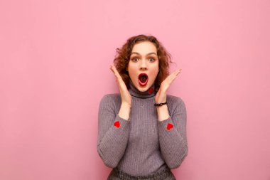 Pembe arka planda kıvırcık saçlı, çekici ve şok olmuş bir kızın yakın plan portresi. Şaşkın bir yüzle kameraya bakıyor ve ellerini kaldırıyor. İzole edilmiş. Boşluğu kopyala
