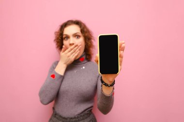 Shocked curly girl covered her mouth with her hand, holding a smartphone with a black screen in her hand, looking into the camera. Focus on smartphone in hand isolated on pink background.