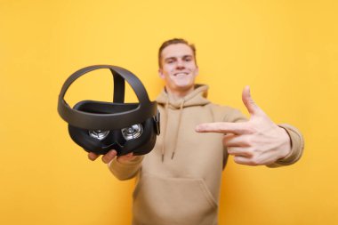 Young man holds a virtual reality helmet in his hand, points a finger at him and smiles. Happy guy shows his finger on VR helmet in his hand on a yellow background.. Focus on VR gadget.