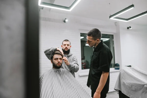 Два перукаря створюють зачіску для бородатого чоловіка в сучасному перукарні. Чоловіча модель вирізала 2 перукарів. Майстер-клас з чоловічих стрижок у перукарні, підготовка перукарської зачіски — стокове фото
