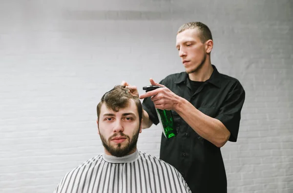 Barber starts to cut the shaggy client, moisturizes the hair with a spray and combs. Portrait of client in hairdresser and professional hairdresser on white wall background, model looking into camera. — ストック写真