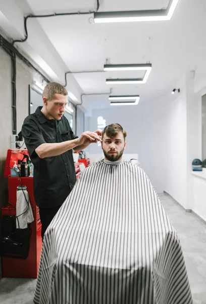 Вертикальний портрет перукаря і його клієнта в яскравому сучасному перукарні. Перукар розрізає ножиці бородатого чоловіка. Створення концепції чоловічої стрижки — стокове фото