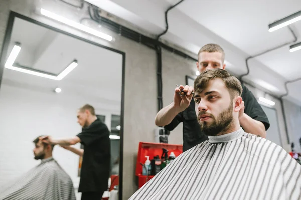 Портрет людини, що сидить у перукарні у кріслі та стриже волосся у перукарні. Чоловічий перукар розрізає бородатого клієнта в сучасному легкому перукарні. Створення чоловічої зачіски . — стокове фото