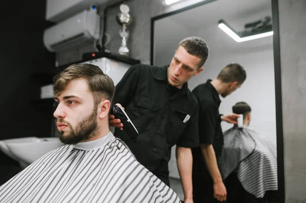 Barber cuts the client with a clipper in his hand in a modern barbershop. Male hairdresser creates hairstyle for handsome bearded man. Barber shop concept. — Stockfoto