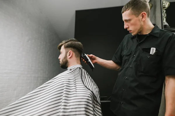 Il barbiere taglia il tagliacapelli del cliente in un parrucchiere da uomo. Parrucchiere crea acconciatura elegante per il cliente barbuto. Uomo che fa taglio di capelli alla moda in barbiere — Foto Stock
