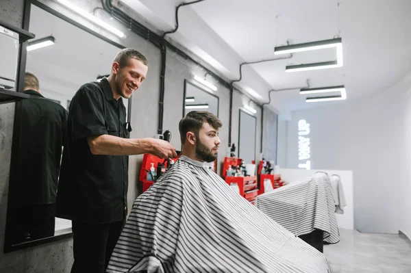 A smiling barber cuts a customer with a clipper in a bright modern barber shop. Happy male hairdresser creates hairstyle to bearded man. Positive barber uses clipper. — Stockfoto