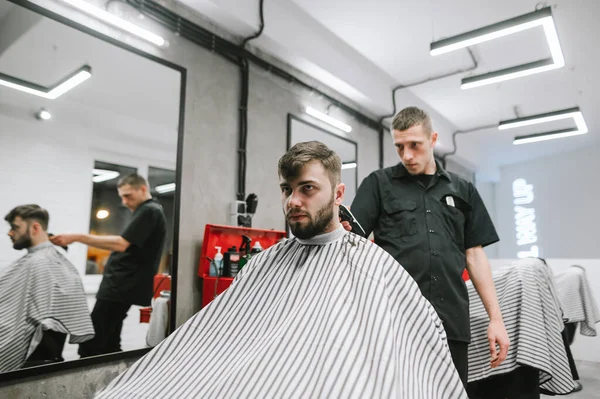 Der männliche Friseur schneidet einen ernstzunehmenden Kunden, der Friseur benutzt einen Clipper. Porträt eines Friseurs schneidet einen Mann mit einem Haarschneider in der Hand, blickt auf seine Haare. Haarschnitt im Friseurladen — Stockfoto