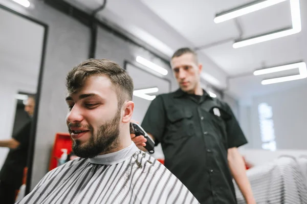 Portrait of happy bearded male client cutting hair in barber. Positive man makes a haircut in a barbershop. Professional barber cuts a cheerful client. — Stockfoto