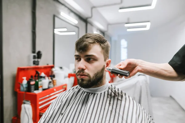 Πορτρέτο ενός γενειοφόρου άντρα που κόβει μαλλιά σε κομμωτήριο ανδρών με σοβαρό πρόσωπο. Ο πελάτης κουρεύει γενειάδα και χτένισμα σε κουρείο, κοντινή φωτογραφία. — Φωτογραφία Αρχείου