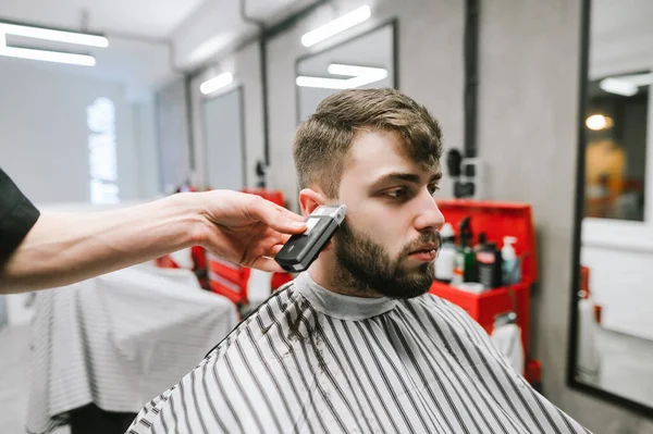 De hand van de kapper snijdt de baardtrimmer van de man door. Portret van een man met een baard die in een kapperszaak zit en kapsel doet. Een mannelijk kapsel maken in een mannenkapsalon. — Stockfoto