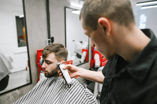 O barbeiro profissional corta o cabelo de um homem barbudo num cabeleireiro moderno e leve. Cabeleireiro masculino com clipper em mãos cria penteado para o cliente. Conceito de barbearia . — Fotografia de Stock
