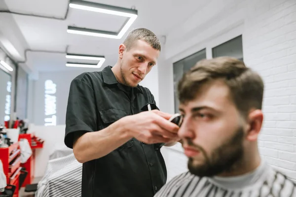 O barbeiro profissional corta um cliente barbudo em uma barbearia leve, olha para o cabelo e usa um cortador de cabelo. Cabeleireiro masculino positivo faz corte de cabelo masculino com um cortador de cabelo. Conceito de barbearia — Fotografia de Stock