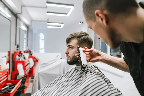 Sitzt ein Mann auf einem Stuhl in einem Friseursalon und schneidet sich mit einem professionellen Friseur die Haare. Friseur trimmt Kundin mit Bartschneider Stilvolle Männerfrisur im Friseursalon. — Stockfoto