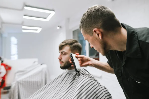 Professional male hairdresser cuts bearded man in modern barbershop, uses clipper. Focusing on a barber with a clipper creates a stylish hairstyle for the client. — Stockfoto
