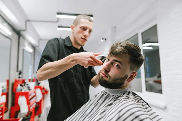 A fun client cuts a professional male hairdresser, sits on a hairdresser and looks into the camera. Barber cuts a cheerful bearded client. Barber shop concept. — Stockfoto