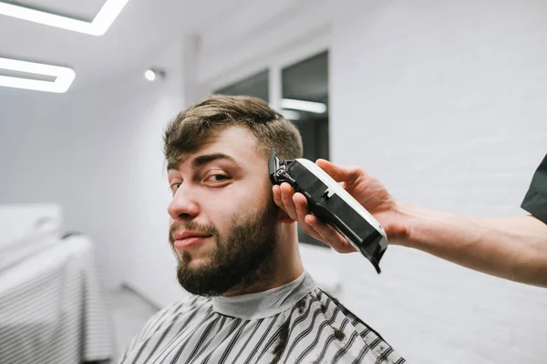 Un ritratto ravvicinato di un uomo barbuto allegro siede su una sedia e un negozio di barbiere e taglia un parrucchiere, un barbiere taglia un clipper.Funny cliente taglia i capelli in un parrucchiere e guarda nella fotocamera — Foto Stock