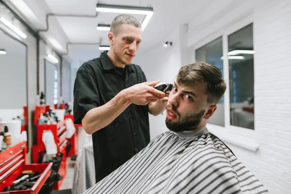 A positive barber cuts a funny client, sits in a chair, looks at the camera and makes a funny face. Dissatisfied client sits in a barber's chair and waits for the haircut to end. — Stockfoto