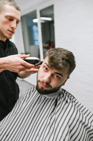 Homem com barba visita uma barbearia, um barbeiro repete criando um penteado com um clipper nas mãos. Cabeleireiro profissional faz penteado elegante para um belo cliente em um cabeleireiro masculino — Fotografia de Stock