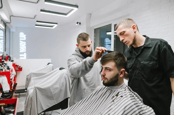 Twee kappers snijden een man met een baard in een licht moderne kapperszaak. Kapsalon leraar leert student kapsels te doen. Kapper Workshop. Barber shop concept. — Stockfoto