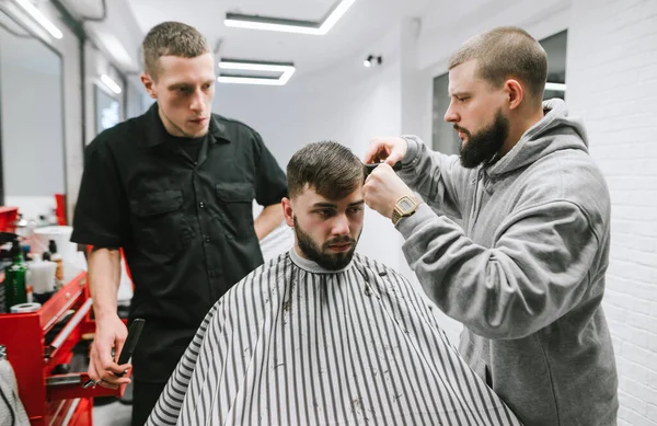 Friseurladen. Ein professioneller Friseur bringt einem Studenten bei, in einem Männerladen ein Modell zu schneiden. Zwei Männer schneiden ein bärtiges Model in einem Friseursalon. Workshop über Männerfrisuren. — Stockfoto