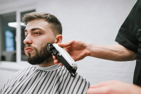 Barbeiro corta a barba de um homem bonito com um cortador. Retrato de perto do cliente da barbearia. Barbeiro correção da barba do cliente no cabeleireiro masculino . — Fotografia de Stock