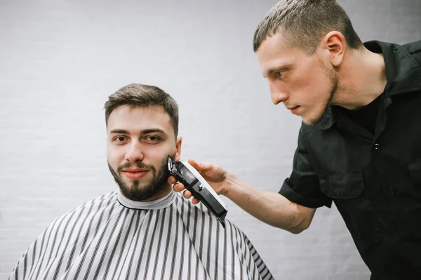 Peluquero profesional corta a un cliente barbudo positivo con un clipper contra una pared blanca, un hombre mira a la cámara y sonríe, un peluquero utiliza un clipper . — Foto de Stock