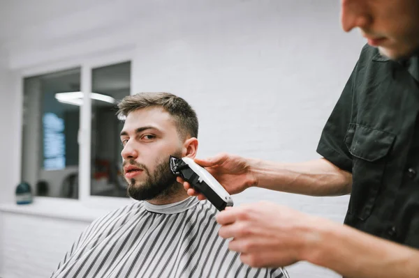 Professional barber makes a beard haircut for a client with a clipper. Hairdresser makes beard correction to handsome man. Man does a haircut at a men's hairdresser and looks into the camera. — Stockfoto