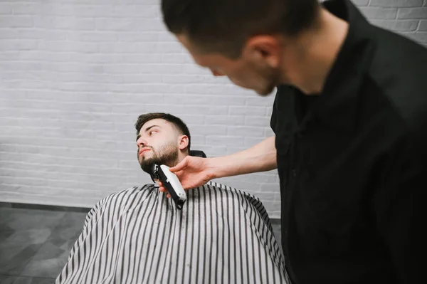 Barber cuts the barber shop's beard clipper. Man deals with the correction of the beard in the men's shop. Hairdresser does a beard hairstyle in a man lying on a chair and looking away — Stockfoto