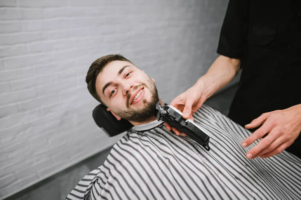 Sonriente hombre guapo hace un corte de pelo de barba, se encuentra en una silla y mira a la cámara con una sonrisa en la cara. Peluquería masculina ajusta la barba del cliente con un cortador. Cliente de barbería feliz — Foto de Stock