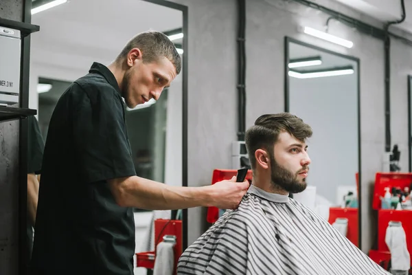 Портрет бородатого клієнта, який ріже волосся на перукарні в чоловічому перукарні, тример для шиї з серйозним обличчям. Чоловічий перукар робить стильну зачіску для бородатого чоловіка — стокове фото