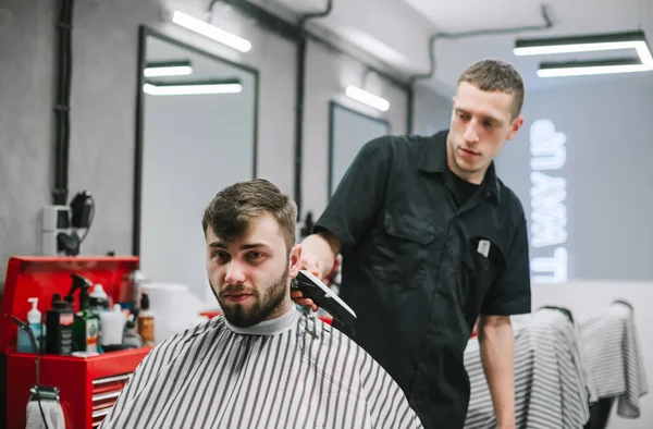 Professional male hairdresser does hairstyle to bearded man in barber shop, client looks into camera with serious face. Barber cuts a man into a client at a men\'s hairdresser