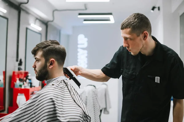 Barber cut client's hair with a clipper in his hands. Barber works in a barbershop , create a stylish hairstyle for a bearded man. Barber shop concept — Stockfoto