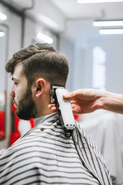 Крупный план руки парикмахера с стрижкой, создает стильную прическу для бородатого мужчины в парикмахерской для мужчин. Парикмахер режет клиента в парикмахерской, закрывается. Вертикальное фото — стоковое фото