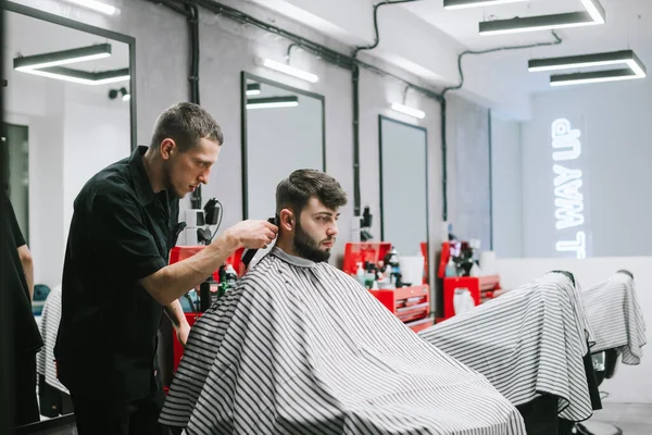 Kapper knipt haar van een bebaarde man met een tondeuse in een stijlvolle herenkapper met een licht interieur. Kapsalon creëert kapsel voor klant in kapperszaak. — Stockfoto