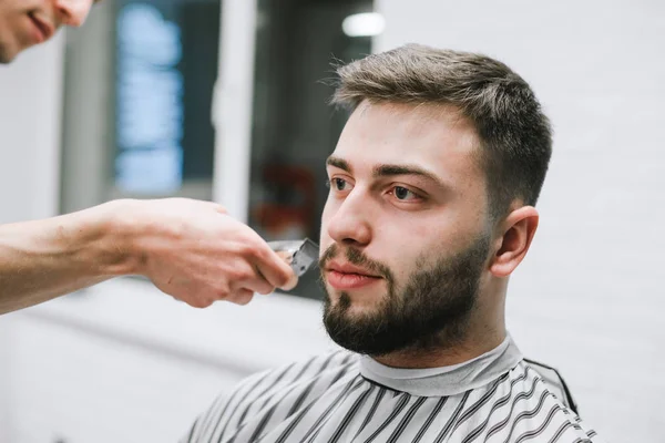 Adult male model with beard trimming at barber shop. Close up portrait of a client doing hairstyle in light barbershop. Male hairdresser creates a stylish hairstyle — Stockfoto