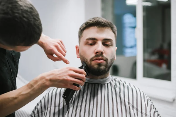 Un parrucchiere professionista maschio fa un taglio di capelli barba in un bell'uomo, usa un clipper. Attraente rende una barba e acconciatura testa in un negozio di barbiere, ritratto primo piano . — Foto Stock