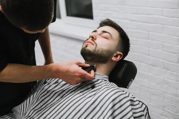 Професійний перукар робить бороду зачіску до клієнта з обрізом у руках, лежачи з закритими очима на стільці в перукарні. Перукар робить з людини бороду, використовуючи обрізку. — стокове фото