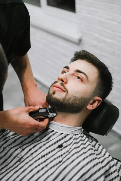 Porträt eines Mannes im Friseursalon, der in einem Stuhl liegt und aufblickt, während sich der Friseur den Bart schneidet. Friseur schneidet dem Kunden ernsthaft seinen Bart. vertikale Nahaufnahme Porträt. — Stockfoto