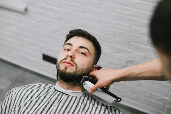 Porträt eines gutaussehenden Mannes, der in einem Friseurstuhl liegt und mit ernstem Gesicht in die Kamera blickt. Friseur schneidet Bart mit einem Clipper in der Client. Friseur macht Bartfrisur inman. — Stockfoto