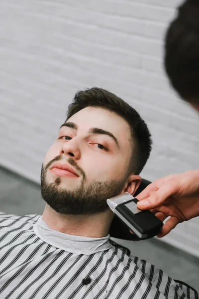 Портрет чоловіка, який лежить в кріслі і дивиться в камеру, перукар, який робить зачіску бороду клієнту, використовує тример. Позитивний перукар розрізає бороду чоловікові з серйозним обличчям. Вертикальний. Барбершоп — стокове фото
