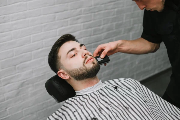 Professional barber makes a beard trimming a trimmer in his hands.Man with a beard is in a chair with a serious face and uses the barbershop.Barber cuts the beard of the client