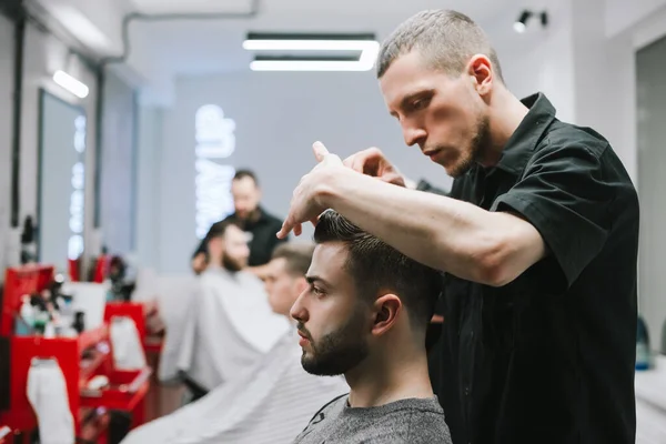 The concubine barber finishes the work of cutting a bearded man, invests hair with gel hands. Male hairdresser styling client's hair after haircut. Barbershop concept. — Stockfoto