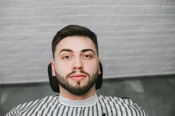 Retrato de un hombre positivo acostado en una silla en una barbería, mirando a la cámara y sonriendo. Alegre cliente en la silla de un peluquero masculino. Cierra la foto. Servicios de peluquería . — Foto de Stock