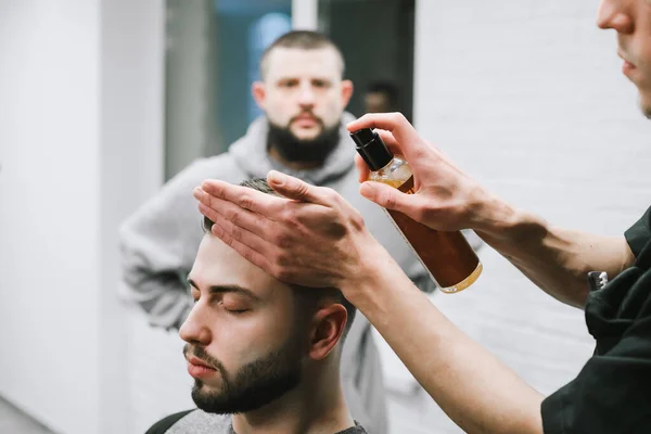 Barbert uses a hair spray when styling a client's hairstyle. Male hairdresser invests hair of bearded client. Barber finishes work on her hairstyle. Hands in focus close up. Barbershop. — Stockfoto