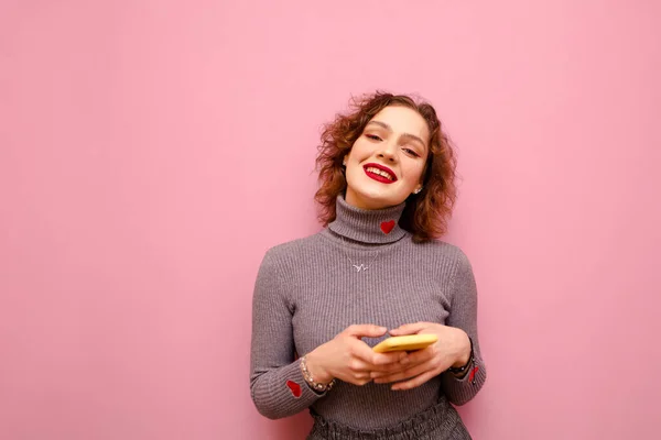 Щаслива кучерява дівчина в сірому светрі стоїть на рожевому тлі зі смартфоном в руці, дивиться в камеру і посміхається. Мила дама з яскравим макіяжем посміхається використовує смартфон ізольовано . — стокове фото