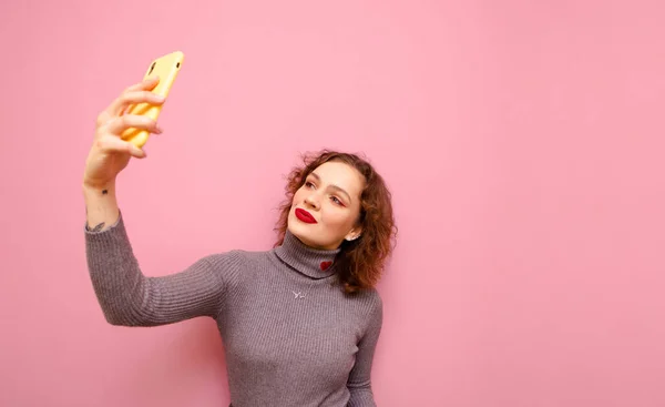 Cute lady with makeup and curly red hair takes selfie on smartphone on pink background, wears gray sweater. Attractive teen girl with red lips posing on smartphone camera, isolated. Copy space — ストック写真