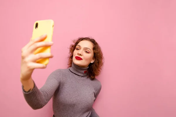 Attractive lady in casual clothes makes selfie on pink background, holds smartphone, wears gray sweater. Lady with red curly hair poses on a camera with a serious face. Isolated. Copy space